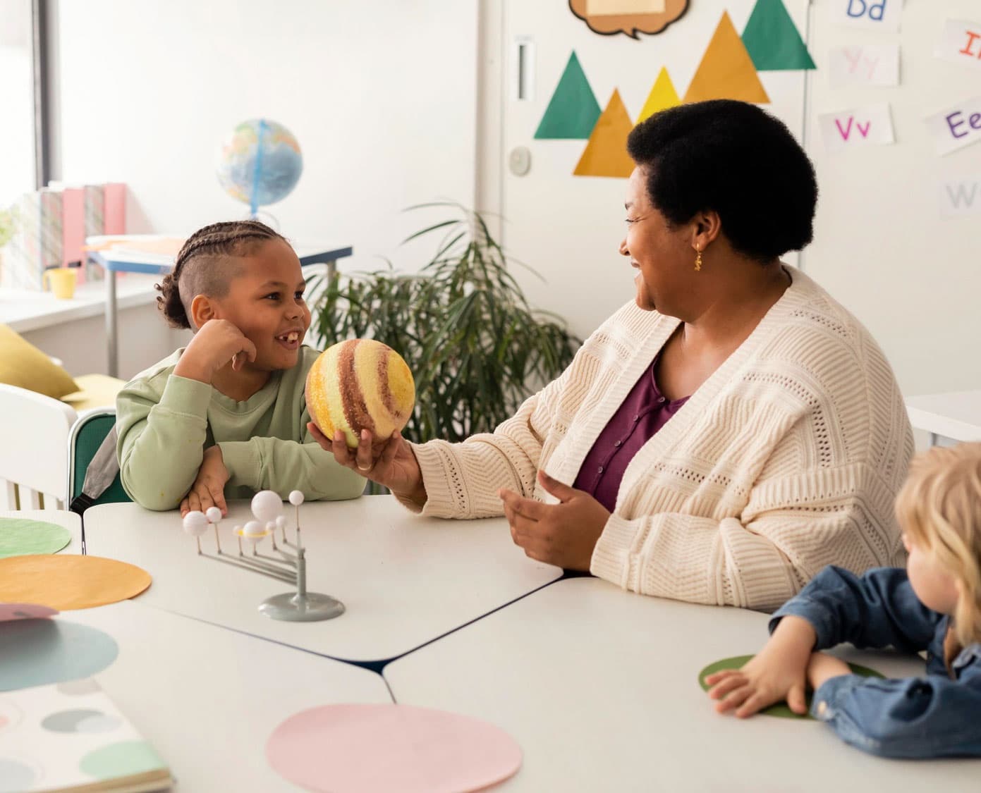 A teacher showing a smiling child a model planet beside a solar system model on the table